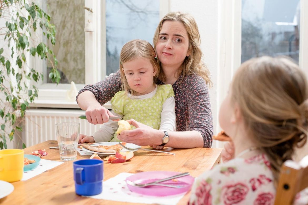 - Copyright: Deutsches Müttergenesungswerk Eine Mutter sitzt mit 2 Kindern am Frühstückstisch und beschmiert ein Brötchen - Copyright: Deutsches Müttergenesungswerk