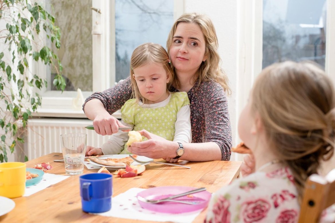Eine Mutter sitzt mit 2 Kindern am Frühstückstisch und beschmiert ein Brötchen