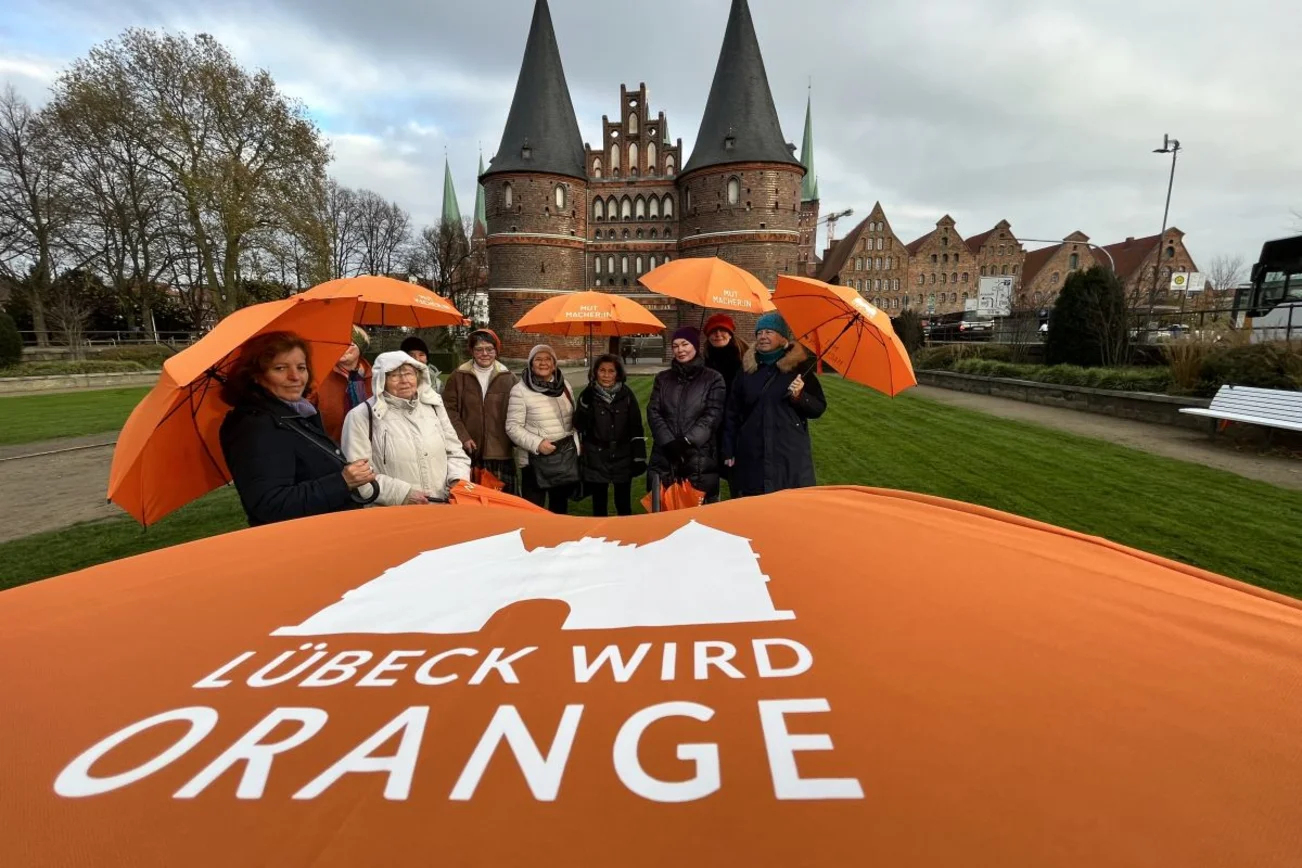 Schriftzug Lübeck wird orange, dahinter Frauen mit orangefarbenen Regenschirmen