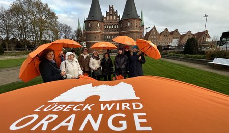 Schriftzug Lübeck wird orange, dahinter Frauen mit orangefarbenen Regenschirmen
