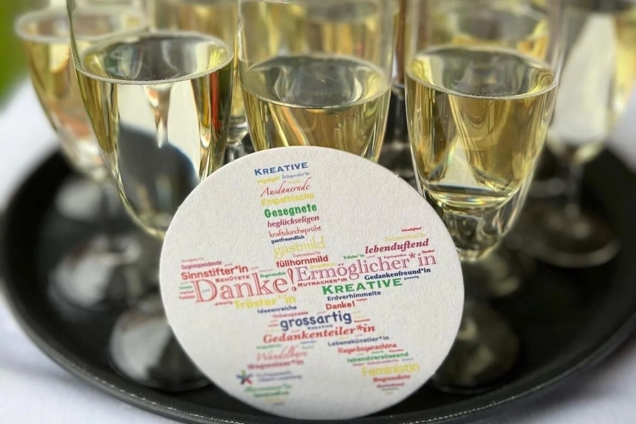 Plakette mit Wörterwolke, dahinter stehen Sektgläser