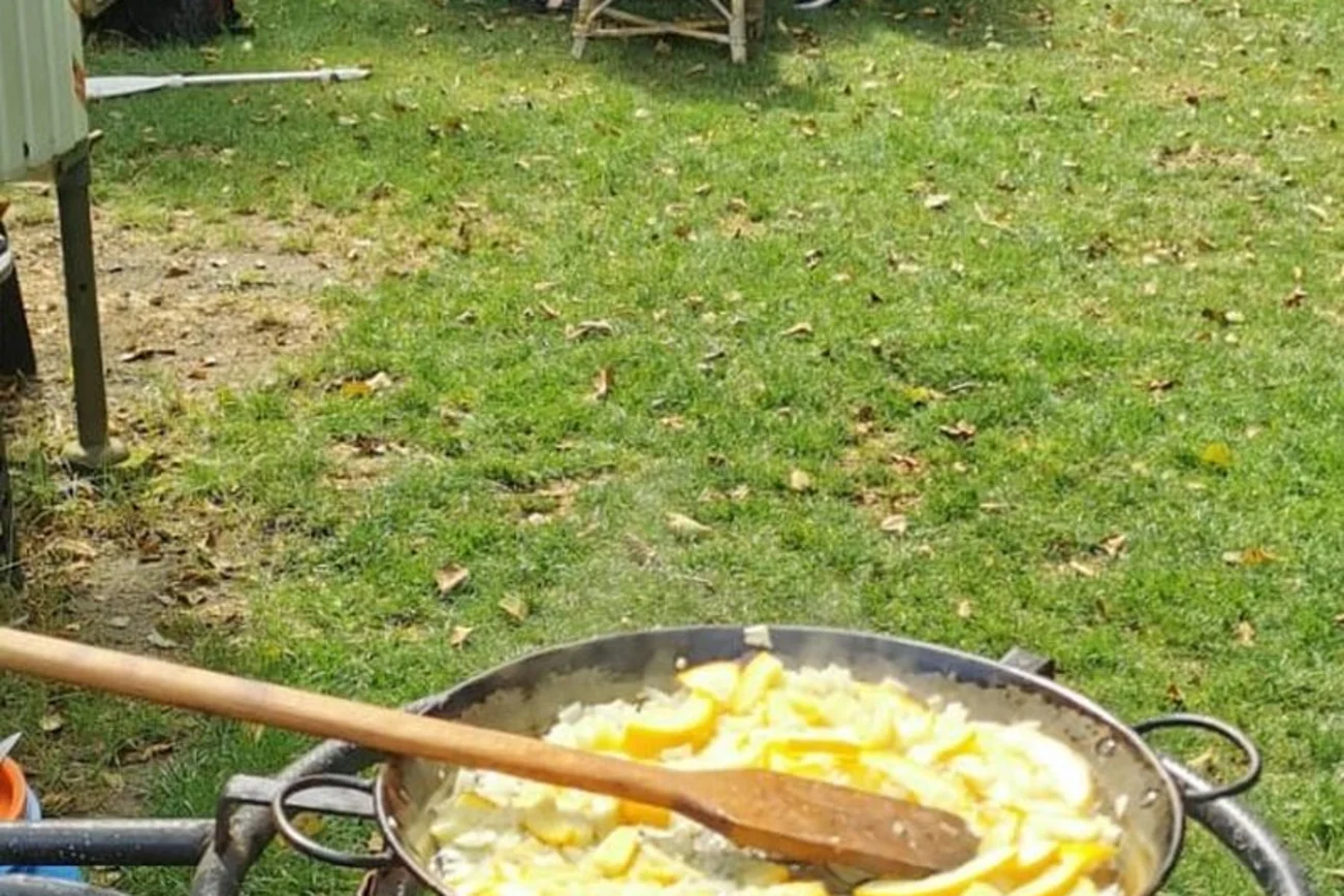 Im Hintergrund sitzen Frauen am Tisch, vorne Topf mit Essen auf Feuer